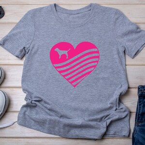 May include: A gray t-shirt featuring a large pink heart design. Inside the heart is a silhouette of a dog and horizontal stripes. The shirt is made of a soft material and has short sleeves.