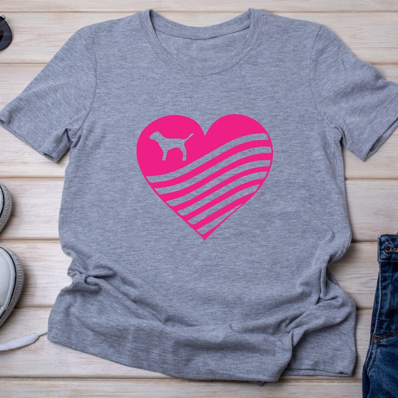 May include: A gray t-shirt featuring a large pink heart design. Inside the heart is a silhouette of a dog and horizontal stripes. The shirt is made of a soft material and has short sleeves.