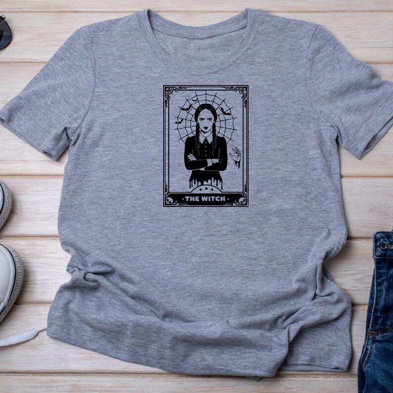 May include: A gray t-shirt featuring a black and white graphic of a woman in a gothic style, framed by a spiderweb and bats. The text "THE WITCH" is at the bottom of the graphic. The shirt is made of a soft material.