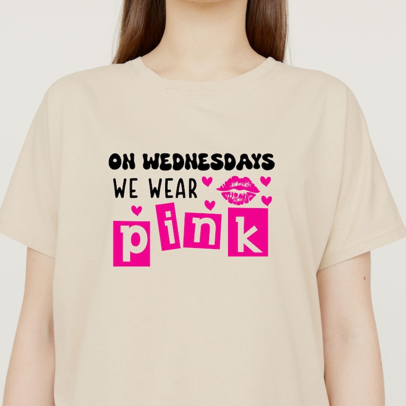 以下が含まれることがあります： ベージュのTシャツに、黒い文字で「On Wednesdays We Wear Pink」と書いてあります。「pink」という単語はピンクの文字で書かれており、ピンクの口紅のグラフィックとハートが描かれています。