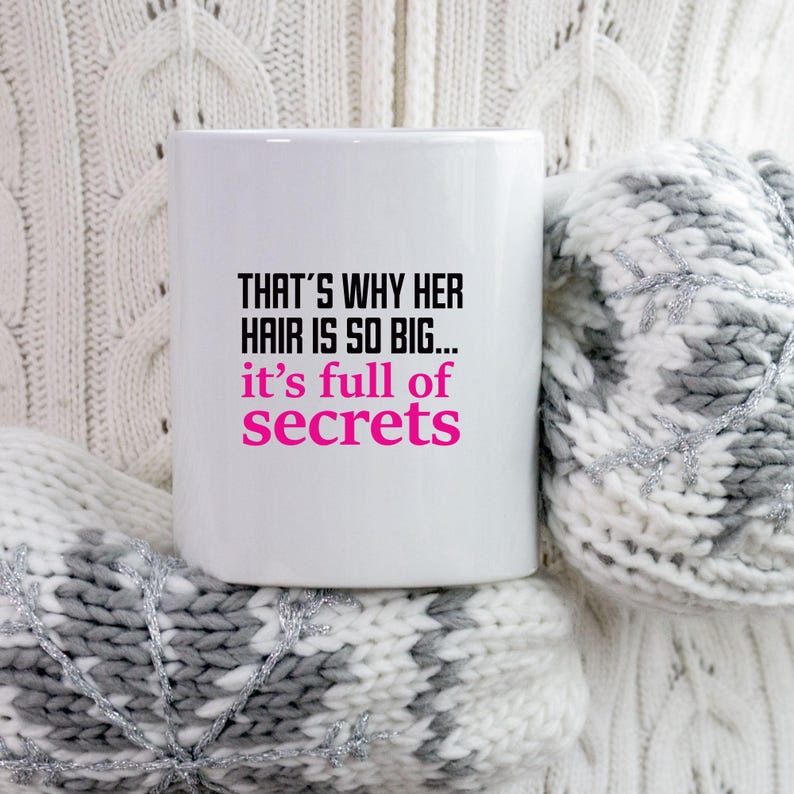 May include: White ceramic mug with the text "THAT'S WHY HER HAIR IS SO BIG... it's full of secrets" in black and pink. The mug is set against a cozy, white and gray knitted background.