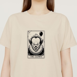 May include: Beige t-shirt featuring a black and white graphic of a clown tarot card. The card depicts a sinister clown face with a balloon and the text "THE CLOWN". The shirt is a neutral color.