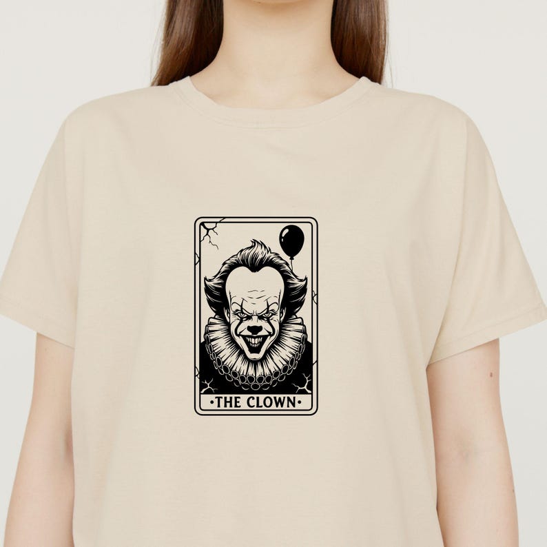 May include: Beige t-shirt featuring a black and white graphic of a clown tarot card. The card depicts a sinister clown face with a balloon and the text "THE CLOWN". The shirt is a neutral color.