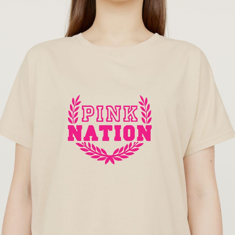 May include: A beige t-shirt with the text "PINK NATION" in bright pink, framed by a decorative wreath. The shirt has short sleeves and a crew neck.
