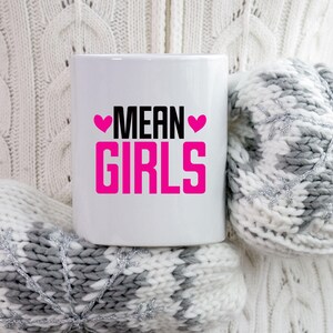 以下が含まれることがあります： 白地に黒とピンクの文字で「MEAN GIRLS」と書かれた白いセラミックマグカップ。テキストは2つのピンクのハートで囲まれています。