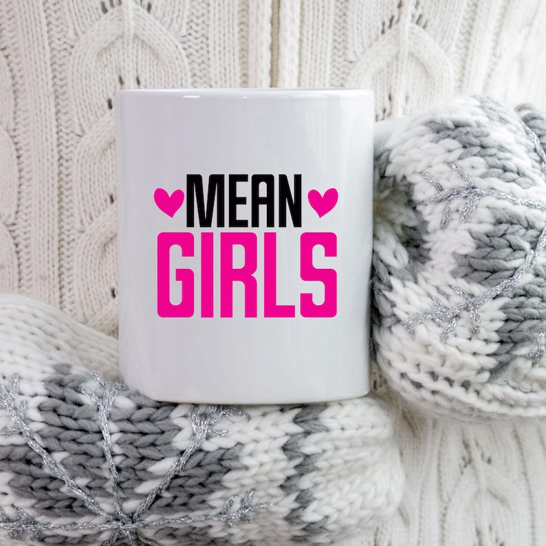 以下が含まれることがあります： 白地に黒とピンクの文字で「MEAN GIRLS」と書かれた白いセラミックマグカップ。テキストは2つのピンクのハートで囲まれています。