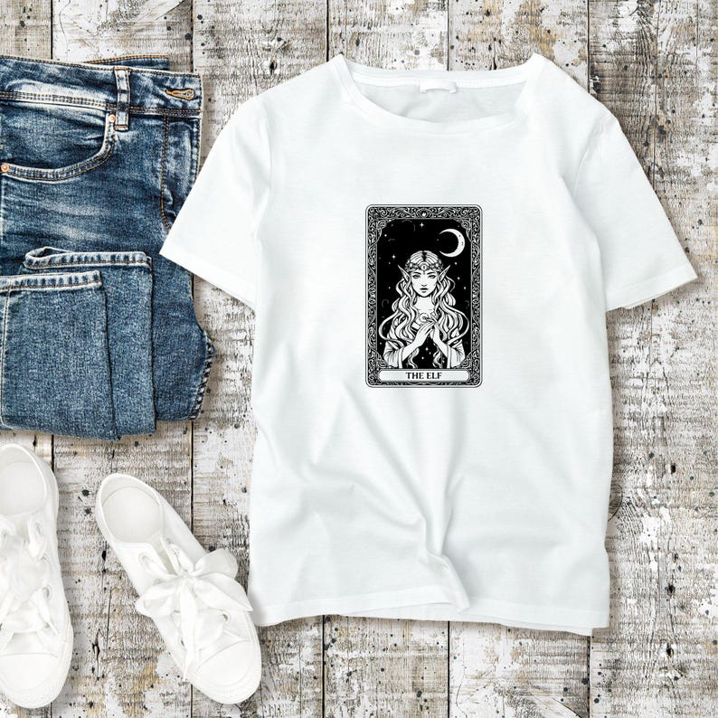 May include: White t-shirt featuring a black and white illustration of an elf tarot card. The design includes an elf with long hair, a crescent moon, and the text "THE ELF". The shirt is paired with blue jeans and white sneakers.