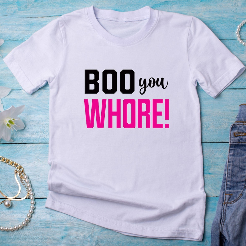 以下が含まれることがあります： 黒い文字で「BOO you WHORE!」と書いてあるピンクの文字の白いTシャツです。