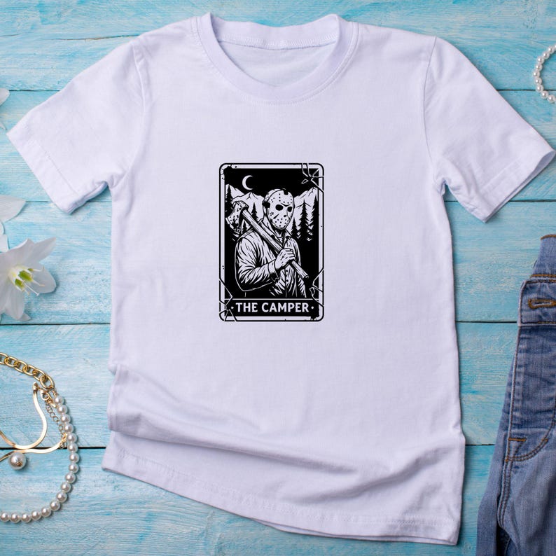 May include: White t-shirt featuring a black and white graphic of a masked figure holding an axe, with the text "THE CAMPER." The design includes a mountain and moon background. The shirt is made of a soft material.
