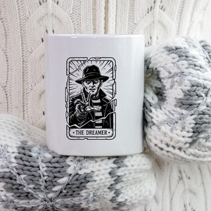 May include: White ceramic mug featuring a black and white illustration of a character in a hat and striped sweater, with the text "THE DREAMER". The design is framed by a cracked border.