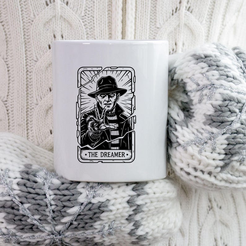 May include: White ceramic mug featuring a black and white illustration of a character in a hat and striped sweater, with the text "THE DREAMER". The design is framed by a cracked border.
