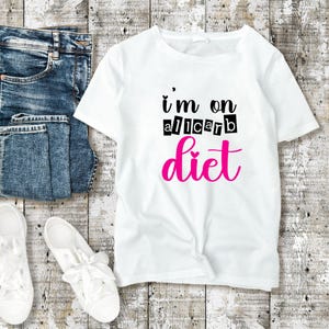 以下が含まれることがあります： 白いTシャツに黒い文字で「i'm on a all carb diet」とピンクのフォントで書かれています。