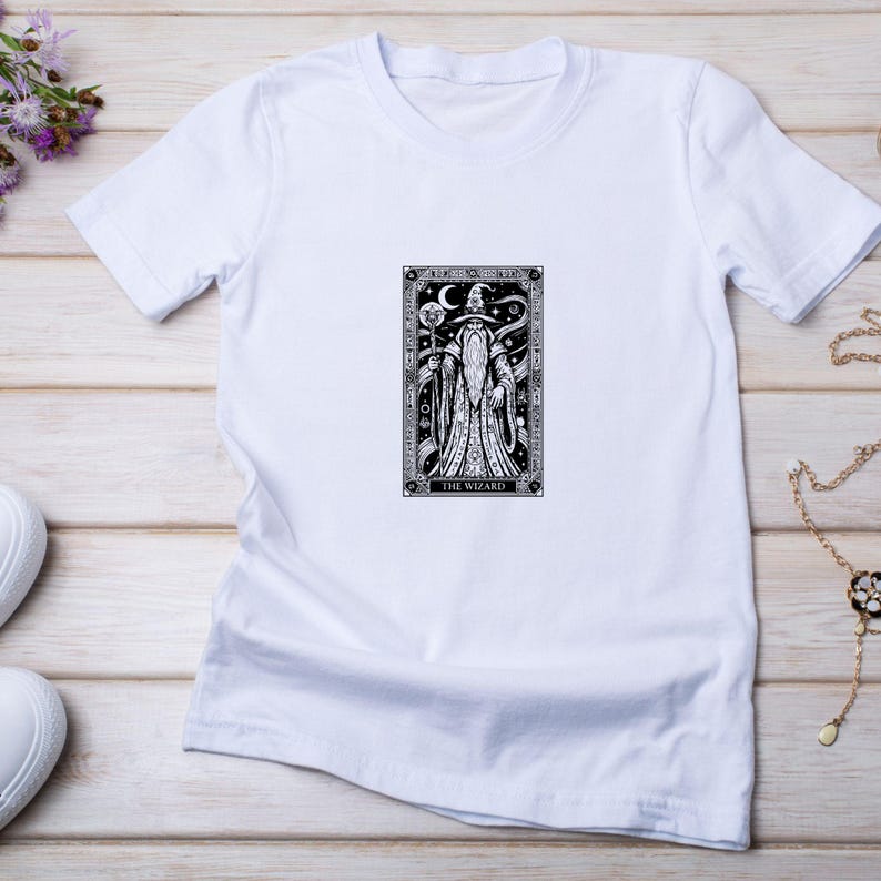 May include: White t-shirt featuring a black and white illustration of a wizard with a staff and crescent moon. The text "THE WIZARD" is at the bottom of the design. The shirt is on a light wood surface.