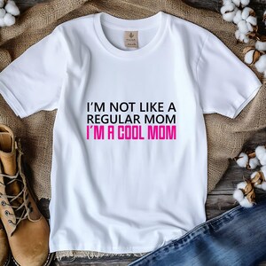 May include: White t-shirt with the text "I'M NOT LIKE A REGULAR MOM I'M A COOL MOM" in black and pink. The shirt is made of a soft material and has a crew neck. The shirt is displayed on a wooden surface.