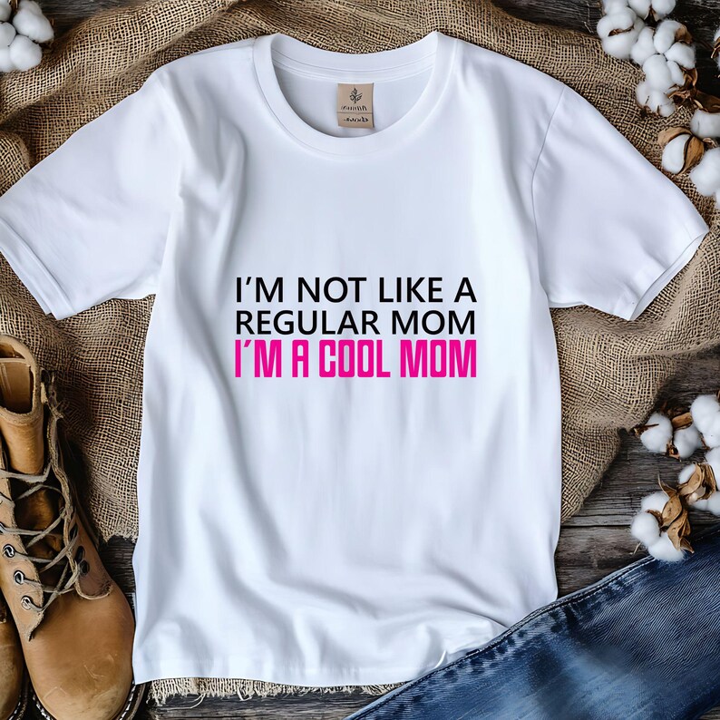 May include: White t-shirt with the text "I'M NOT LIKE A REGULAR MOM I'M A COOL MOM" in black and pink. The shirt is made of a soft material and has a crew neck. The shirt is displayed on a wooden surface.