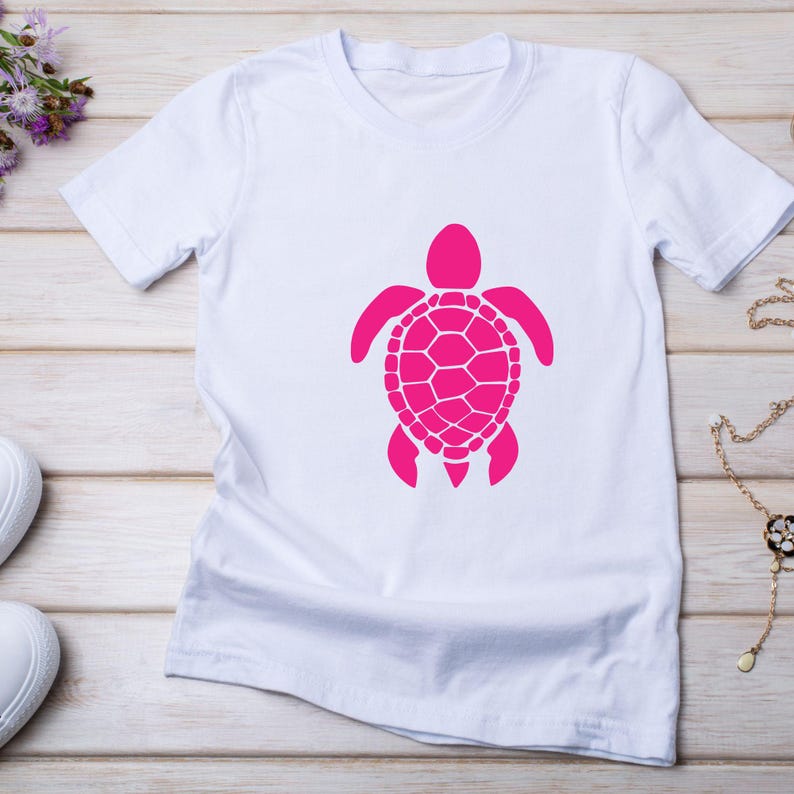 May include: White t-shirt featuring a bright pink sea turtle graphic. The turtle design is centered on the shirt and has a detailed shell pattern. The shirt is made of a soft material and is displayed on a light wood surface.