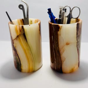May include: Two marble pencil holders with different patterns. One holder has a green, yellow, and brown pattern. The other holder has a brown, white, and orange pattern. Both holders are filled with pens, pencils, and scissors.