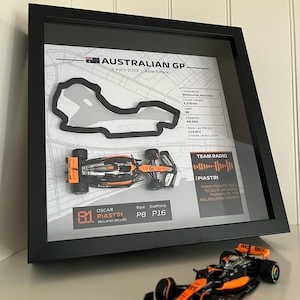 May include: A framed display featuring a 3D map of the Australian GP circuit, a miniature McLaren MCL60 race car, and text detailing race statistics, Oscar Piastri's name, and a team radio quote. A matching miniature race car sits in front.