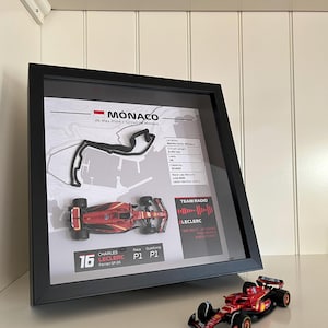 May include: A framed display featuring a 3D Monaco Grand Prix circuit map, a miniature red Ferrari SF-24 race car, and race statistics for Charles Leclerc. A matching miniature red Ferrari race car sits on the surface in front of the black frame.