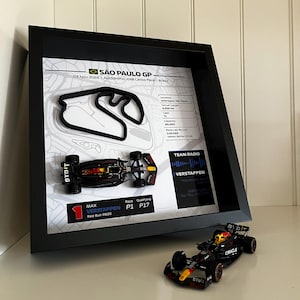 Könnte beinhalten: Ein schwarzer Rahmen mit einer 3D-Streckenkarte des GP von São Paulo, einem Miniatur-Rennwagen Red Bull RB20 und Textdetails, einschließlich des P1-Rennergebnisses von Max Verstappen. Ein passender Miniatur-Rennwagen steht vor dem Rahmen.