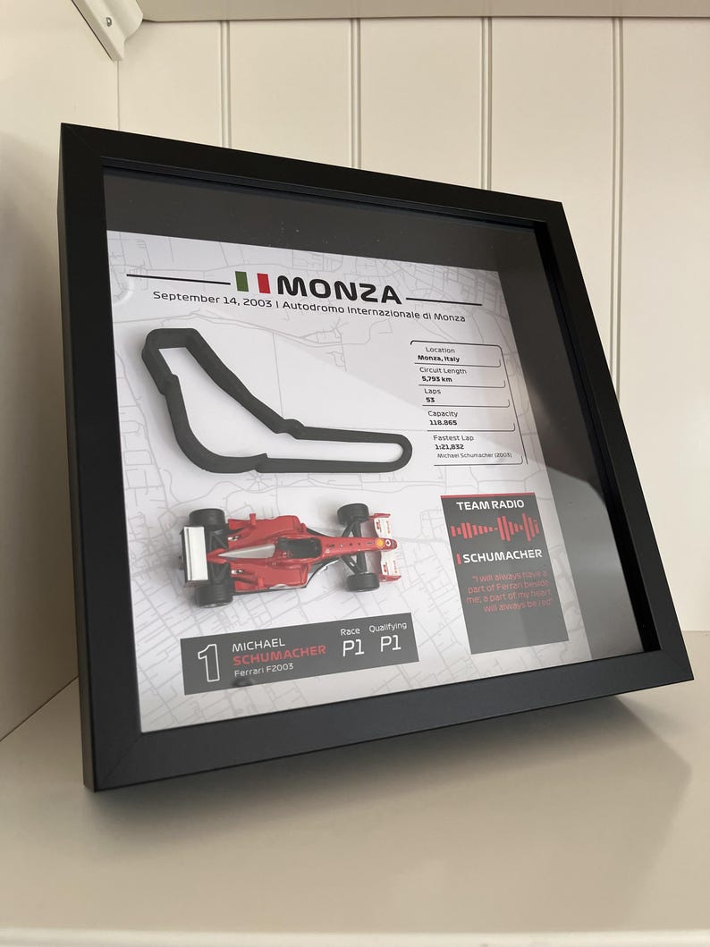 May include: A framed display featuring a 3D black outline of the Monza race track, a miniature red Ferrari F2003 race car, and text detailing the September 14, 2003 race, including circuit length, lap count, and Michael Schumacher's P1 qualifying result.