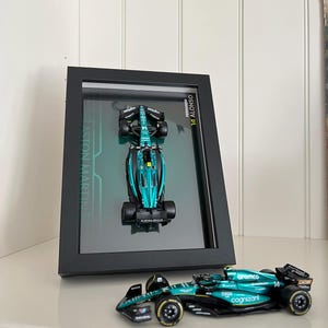 Peut inclure: Un modèle réduit d'une voiture de course Aston Martin de Formule 1 encadré dans un coffret noir. La voiture est de couleur vert d'eau et noire avec des logos de sponsors. Un autre modèle identique est posé devant le cadre.