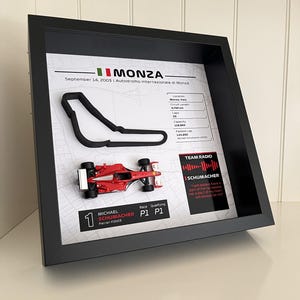 Könnte beinhalten: Ein schwarzer Rahmen mit einem schwarzen 3D-Umriss der Rennstrecke Monza, einem roten Miniatur-Rennwagen Ferrari F2003 und gedrucktem Text über das Rennen vom 14. September 2003, einschließlich Streckenstatistiken und einem Zitat von Michael Schumacher.