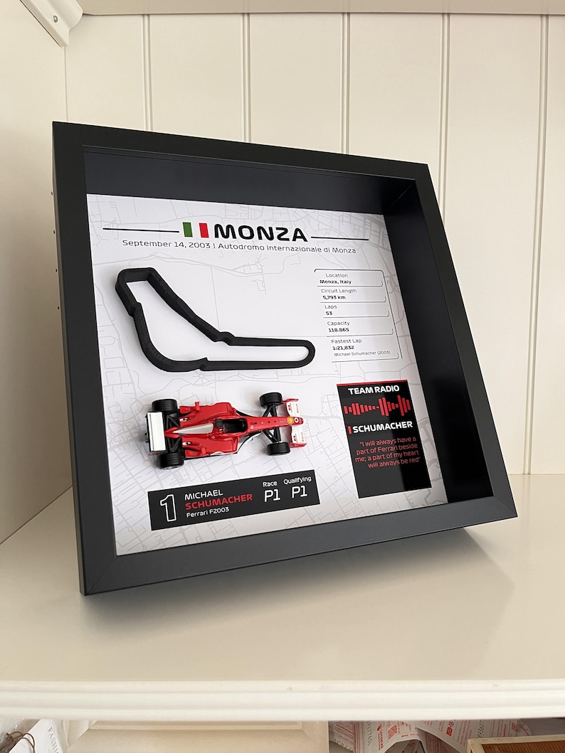 May include: A black framed display featuring a 3D black outline of the Monza race track, a miniature red Ferrari F2003 race car, and printed text detailing the September 14, 2003 race, including track statistics and a quote from Michael Schumacher.