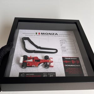 May include: A framed display featuring a red miniature Formula 1 race car, a black 3D track outline of Monza, and a printed map background. The display includes text details about the 2003 Italian Grand Prix, Michael Schumacher, and a Team Radio quote.