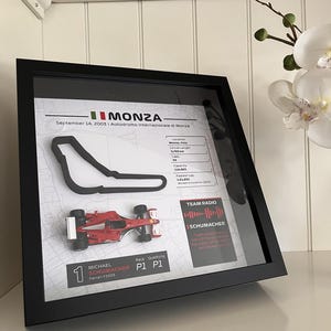 May include: A framed display featuring a black 3D outline of the Monza race track, a miniature red Ferrari F2003 race car, and text detailing the September 14, 2003 race, including circuit stats, a Team Radio sound wave, and Michael Schumacher's P1 results.