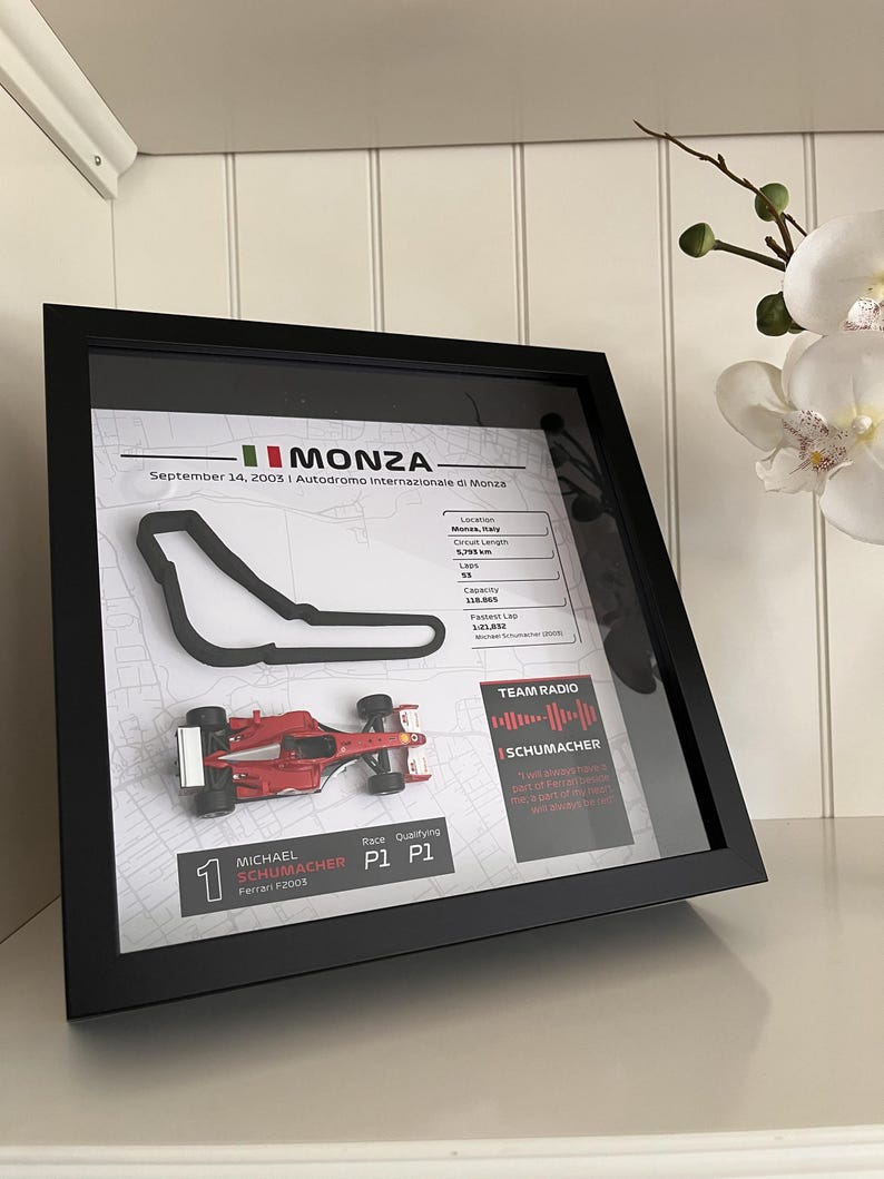 May include: A framed display featuring a black 3D outline of the Monza race track, a miniature red Ferrari F2003 race car, and text detailing the September 14, 2003 race, including circuit stats, a Team Radio sound wave, and Michael Schumacher's P1 results.