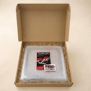 May include: A square brown cardboard shipping box containing a square object wrapped in clear bubble wrap, surrounded by tan paper filler. A black thank you card and a small white sticker with the text LEGACYTRACKS are placed on top.
