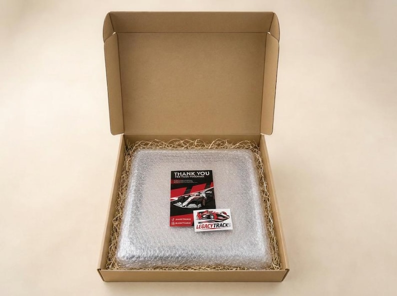 May include: A square brown cardboard shipping box containing a square object wrapped in clear bubble wrap, surrounded by tan paper filler. A black thank you card and a small white sticker with the text LEGACYTRACKS are placed on top.