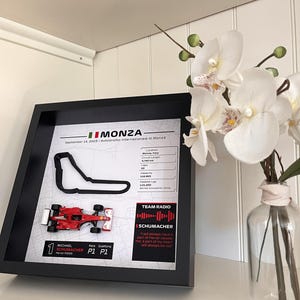 May include: A framed display featuring a black outline of the Monza race track, a miniature red Ferrari F2003 Formula 1 car, and printed statistics including the date September 14, 2003, circuit details, and a quote from Michael Schumacher.