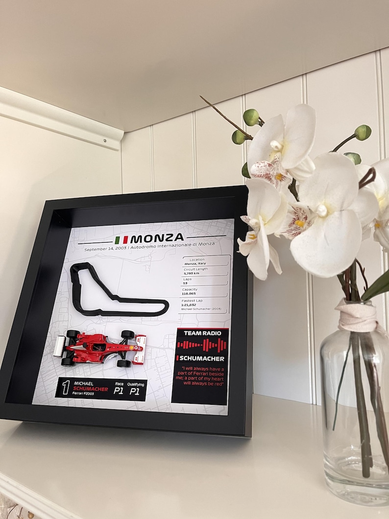 May include: A framed display featuring a black outline of the Monza race track, a miniature red Ferrari F2003 Formula 1 car, and printed statistics including the date September 14, 2003, circuit details, and a quote from Michael Schumacher.