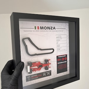 May include: A framed display featuring a black outline of the Monza race track, a miniature red Ferrari F2003 race car, and text detailing the September 14, 2003 race, including circuit statistics, Michael Schumacher's P1 qualifying result, and a team radio quote.
