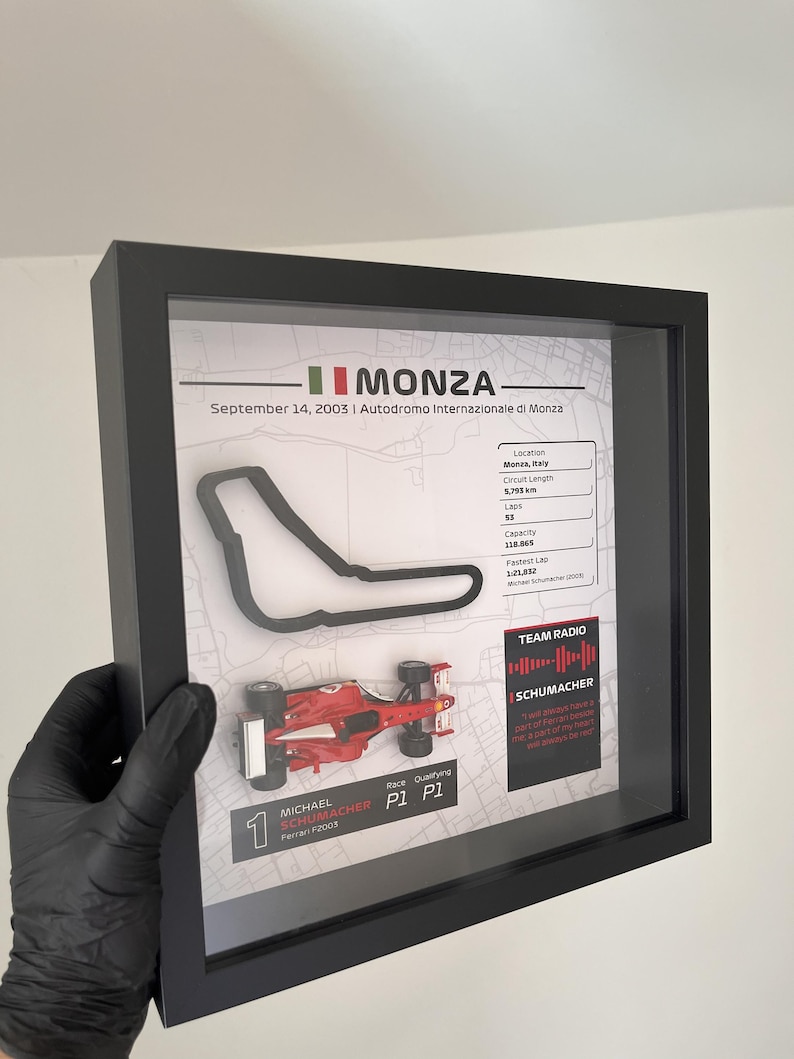 May include: A framed display featuring a black outline of the Monza race track, a miniature red Ferrari F2003 race car, and text detailing the September 14, 2003 race, including circuit statistics, Michael Schumacher's P1 qualifying result, and a team radio quote.
