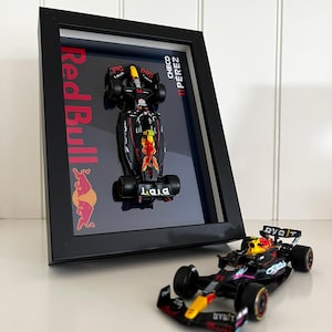 Peut inclure: Un cadre noir présentant un modèle réduit de voiture de Formule 1 Red Bull monté verticalement avec le texte Red Bull et Checo Perez 11, accompagné d'une voiture de course miniature noire, jaune et rouge assortie placée devant le cadre.
