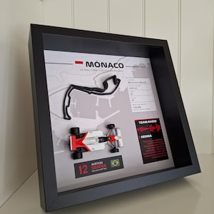 May include: A framed 3D display featuring a black model of the Monaco race track, a miniature white and red McLaren MP4/4 race car, and text detailing the 1988 race, including Ayrton Senna's pole lap record and a Team Radio quote in a black box.