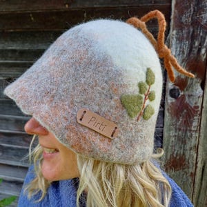 Può includere: Un cappello in lana infeltrita nei toni del bianco, grigio e arancione, con un'etichetta in pelle marrone impressa con il testo Pirti. Il cappello è decorato con un'applicazione a foglia verde e un laccio decorativo in filo arancione.