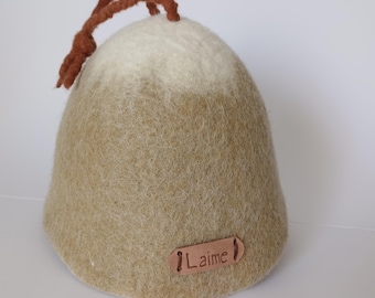 May include: A beige felt sauna hat with a brown drawstring and a leather tag that reads "Laimē". The hat has a rounded shape and a two-tone colour scheme, transitioning from light beige to a darker beige.