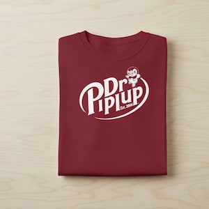 May include: A folded maroon t-shirt with a white graphic. The graphic features the words "Dr. Piplup" in a stylized font, with a cartoon penguin character and the text "Est. 2006". The shirt is on a light wood-grain surface.