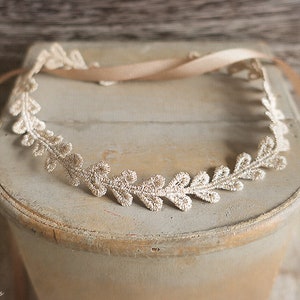 May include: A delicate, cream-colored lace headband with a leaf design. The headband is adorned with a matching ribbon for tying. The piece is displayed on a light-colored, distressed wooden surface, suggesting a vintage or rustic aesthetic.