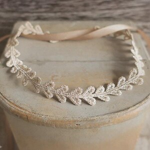 May include: A delicate, cream-colored lace headband with a leaf design. The headband is placed on a light-colored, weathered surface, with a matching ribbon tied at the back. A beautiful accessory for special occasions.