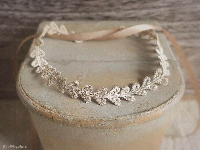 May include: A delicate, cream-colored lace headband with a leaf design. The headband is placed on a light-colored, weathered surface, with a matching ribbon tied at the back. A beautiful accessory for special occasions.