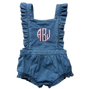 May include: Blue denim baby romper with ruffled straps and hem. The romper features a pink embroidered monogram in the center. The garment is designed with a bubble silhouette.