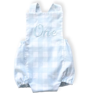 May include: Light blue and white gingham baby romper with the word "One" embroidered in light blue script across the chest. The romper has a bubble shape and cross-back straps.