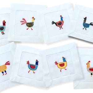 May include: Set of eight white linen cocktail napkins with embroidered colorful rooster designs. Each napkin features a different rooster design with various colors and patterns.