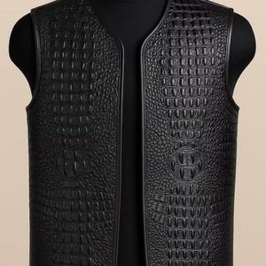 Puede incluir: Un chaleco negro sin mangas que presenta una textura en relieve de piel de cocodrilo en todos los paneles frontales. La prenda tiene un escote sin cuello, bordes lisos de aspecto de cuero negro y un cierre de cremallera central frontal.