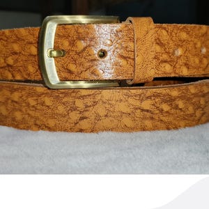 May include: A textured, tan leather belt with a gold-colored buckle. The belt is rolled up and resting on a white surface. The belt has a detailed pattern and a classic design.